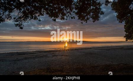Silhouette di persone che scattano foto con il telefono cellulare, il sole che sorge sull'isola di Rangitoto. Incorniciato da alberi di Pohutukawa a Milford Beach, Auckland. Foto Stock