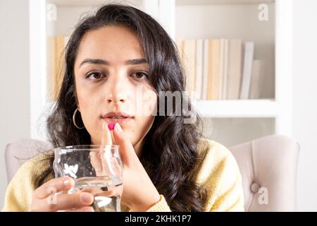 Pillola di tenuta, ritratto di bella giovane donna che tiene la pillola e un bicchiere di acqua dolce. Ragazza a casa in abiti casual. Mal di testa, integratori medicinali Foto Stock