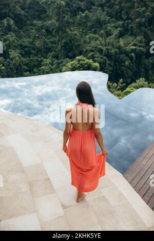 Vista posteriore di una giovane donna che cammina verso la piscina nel resort di lusso. Modello femminile in sundress a bordo piscina. Foto Stock