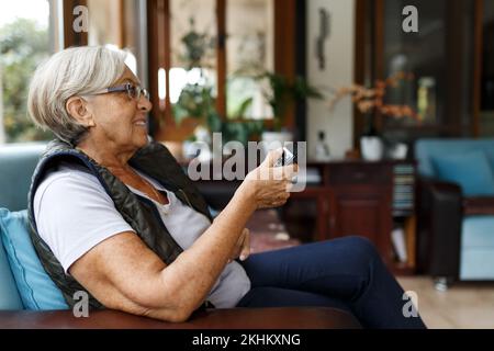 Donna anziana sorridente felice con i capelli grigi seduti sul divano nel soggiorno, puntando un telecomando tv per cambiare canale, entusiasta di guardare Foto Stock