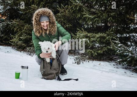 Cura degli animali domestici nella stagione fredda invernale. Donna felice e cane maltese in zaino a piedi, divertirsi e bere tè caldo all'aperto in inverno neve parco natura Foto Stock