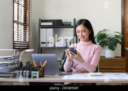 Le giovani donne asiatiche lavorano con documenti finanziari a casa contare su calcolatrice prima di pagare le ricevute fiscali online per telefono. Donna che progetta millennial Foto Stock