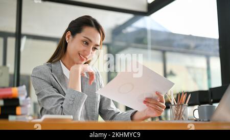 Attraente donna d'affari asiatica millennial che legge le informazioni sui documenti, analizza i dati finanziari, lavora alla sua scrivania. Foto Stock