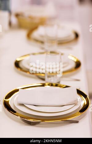 Banchetti, ristorante. Elegante e festoso tavolo minimalista con piatti oro-bianchi, bicchieri, tovaglioli di cotone e posate su tavolo con tovaglia bianca. Foto verticale. Foto Stock