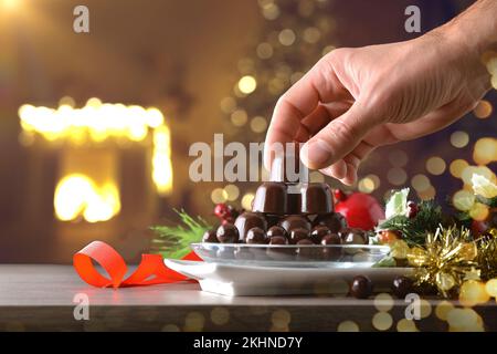 Mano che raccoglie un bonbon di cioccolato fondente da una pila di caramelle al cioccolato su un tavolo di legno con decorazione di Natale e sfondo caldo festa in casa. FR Foto Stock