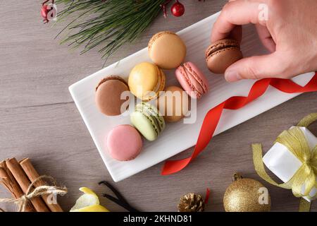 Raccolta a mano di un macaron al cioccolato da un piatto pieno di vari sapori su un tavolo di legno con decorazione natalizia. Vista dall'alto. Foto Stock