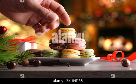 Mano raccogliendo un macaron al cioccolato da un piatto pieno di vari sapori su un tavolo di legno con sfondo natalizio con luci. Vista frontale. Foto Stock