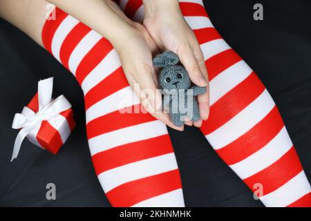 Ragazza in calze natalizie seduta con coniglio lavorato a maglia grigio in mano. Simbolo del Capodanno cinese 2023, abito femminile per la celebrazione Foto Stock