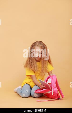 Bambini e tecnologie moderne. Curly studentessa sta mettendo un tablet digitale nel suo zaino rosa, studiando o giocando gioco seduto su sfondo giallo arancione studio, spazio copia gratuito Foto Stock