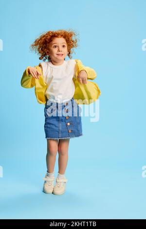 Ritratto di carina bambina, bambino con capelli ricci rossi in posa, saltando isolato su sfondo blu Foto Stock