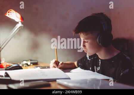 Piccolo ragazzo diligente che fa i compiti mentre ascolta la musica con le cuffie Foto Stock