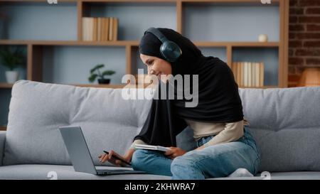 Ragazza studentesca araba islamica donna musulmana indossata nera hijab occupato donna in cuffie con notebook guardare webinar lezione on-line corso di formazione scrivere Foto Stock