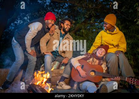Felici amici multirazziali in campeggio ridendo a suonare la chitarra di notte godendo il falò. Foto Stock