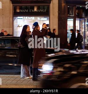 Il turista femminile asiatico scatta una foto su Regent Street in una notte d'inverno, indossando un cappello di lana. Pedoni e automobili passano da sfocato dal movimento. Londra. Foto Stock