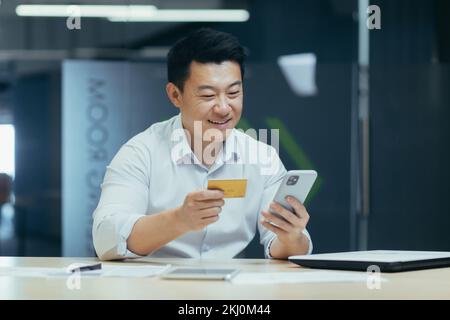 Giovane asiatico seduto alla scrivania in ufficio, paga con carta di credito online, effettua l'ordine per telefono, effettua transazioni con l'app mobile banking. Foto Stock
