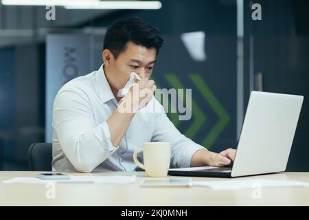 Un giovane freelance asiatico, un lavoratore d'ufficio si è ammalato al lavoro. È seduto al tavolo, ha un naso che cola, gli asciuga il naso con un tovagliolo, si sente male, beve tè caldo, medicina. Foto Stock