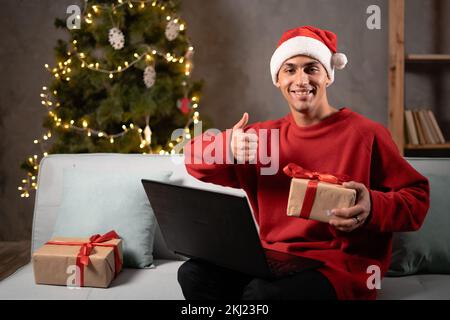 Un uomo arabo entusiasta che mostra un gesto con il pollice in mano e tiene in mano un regalo per videochiamare la famiglia sul laptop di casa. Ragazzo felice indossa il cappello di santa che tiene presen di Natale Foto Stock