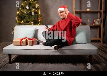 Giovane uomo arabo felice sedersi sul divano a casa tenere telefono videochiamate distanza, Natale online in app mobile utilizzare smartphone videochat chiamata app wavi Foto Stock