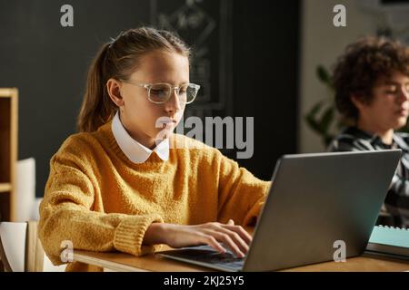 Studentessa di occhiali che si concentra sul suo lavoro online sul computer portatile mentre si siede alla scrivania a lezione a scuola Foto Stock