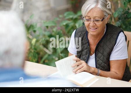 Sposi anziani amorevoli che riposano in giardino e che godono giorni, seduto outdoor.Woman libro di lettura, l'uomo di fronte a lei Foto Stock