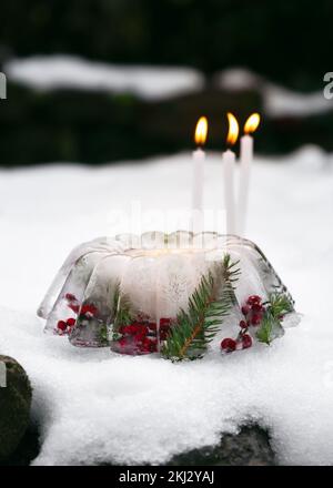 Lanterna di Natale fatta a mano a forma di torta fatta di ghiaccio, bacche di rosa rossa, rami di bacche agrifoglio e ramoscelli di thuja con candele brucianti nella neve. Foto Stock