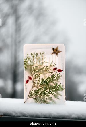 Lanterna di Natale fatta a mano di ghiaccio, bacche di rosa rossa, ramoscelli di thuja e una candela dietro in giardino di sera innevato. Idee facili del mestiere per i capretti Foto Stock