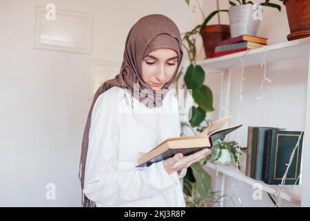 La giovane donna in sciarpa della testa ha letto il libro levandosi in piedi vicino alla libreria. Ragazza teen musulmana che si prepara per gli esami di ingresso. Concetto di istruzione Foto Stock