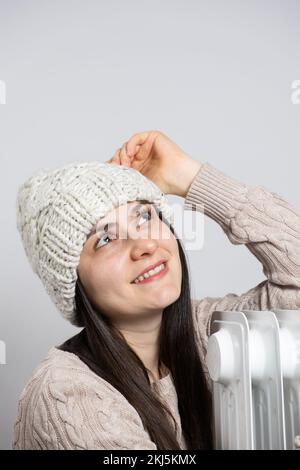 Una donna brunetta in un cappello abbraccia un riscaldatore ad olio, si riscalda in inverno. Foto Stock