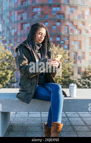 Donna ispanica focalizzata in outerwear messaggi di testo sul cellulare mentre si siede su una panca di pietra con thermos di caffè caldo in città Foto Stock