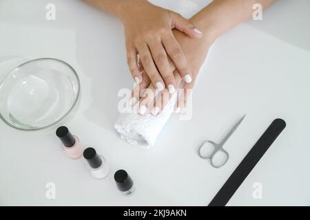 Le mani delle donne ben curate con manicure bianche sono sdraiate sul rullo dell'asciugamano, gli accessori di manicure sono sdraiati sul tavolo vicino ad esso. Primo piano delle mani, vista dall'alto, senza volto Foto Stock