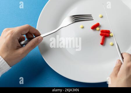 Composizione delle mani che tengono la forchetta e il coltello sopra la piastra con le pillole, su sfondo blu Foto Stock