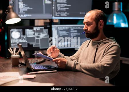 Ingegnere software che analizza il codice sorgente con documenti e computer, prendendo appunti e lavorando allo sviluppo web. Programmatore maschio che confronta i file con i dati del server online dopo ore. Foto Stock