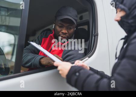 Persona in giacca puffy siglando carte tenuto fuori la finestra del furgone bianco da giovane uomo nero di consegna adulto che indossa il pullover rosso. Scatto orizzontale . Foto di alta qualità Foto Stock