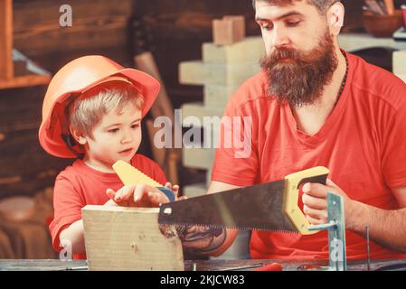 Concetto di giochi educativi. Ragazzo, bambino occupato nel casco protettivo imparare ad usare la sega a mano con papà. Padre, genitore con barba che insegna piccolo figlio a. Foto Stock