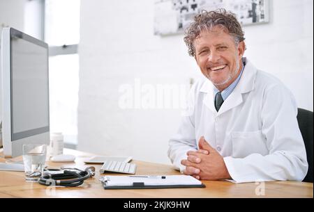 Sempre felice di aiutare. Ritratto di un medico sorridente maturo seduto su una scrivania nel suo ufficio. Foto Stock