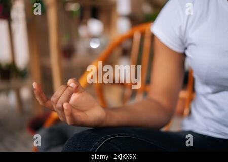 Primo piano ripresa di una serena donna africana irriconoscibile che mette le mani nella postura namaste sul ginocchio seduto in posizione lotus a casa, facendo yoga. Foto Stock