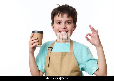 Ritratto isolato sfondo bianco: Felice ragazzo adolescente barista mostra OK segno, vendendo caffè da asporto in tazza di carta ecologica Foto Stock