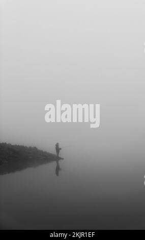 Una scala di grigi verticale di un pescatore in piedi sul bordo di una riva del lago e la pesca in una giornata di nebbia Foto Stock