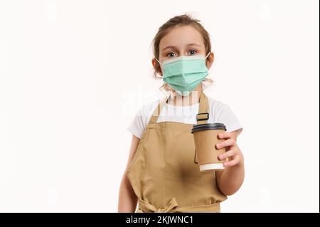 Adorabile bambina in grembiule dello chef e maschera medica protettiva verde, distribuendo una bevanda calda da asporto in tazza di carta Foto Stock