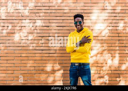 Ritratto di un giovane africano sorridente che indossa una felpa gialla con cappuccio, jeans e occhiali da sole davanti a una parete di mattoni. Il ragazzo sta indossando anelli Foto Stock