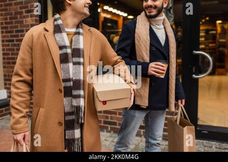 vista parziale di uomini gay eleganti e felici con acquisti e caffè da andare vicino sfocata vetrina sulla strada urbana, immagine stock Foto Stock