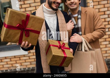 vista ritagliata di una coppia gay felice e alla moda con borse per lo shopping e scatole da regalo sulla strada della città, immagine stock Foto Stock