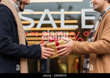 vista parziale degli uomini gay sorridenti che tengono la scatola del regalo vicino alla vetrina sfocata con la scritta di vendita, immagine di scorta Foto Stock