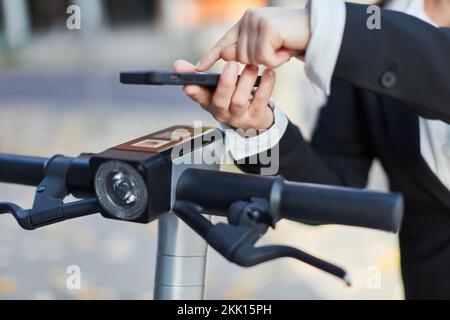 Mano di donna d'affari che noleggia e-scooter con smartphone per le strade Foto Stock