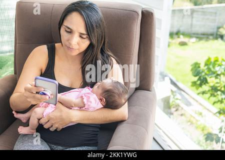 Madre giovane che controlla il suo telefono delle cellule che siede su un divano a casa con il suo bambino vicino ad una finestra con un giardino Foto Stock
