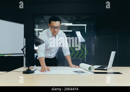 Bel giovane uomo asiatico architetto, designer, ingegnere si pone serio e concentrato in ufficio alla scrivania in una camicia bianca e occhiali, lavora con piani, documenti, disegni. Foto Stock