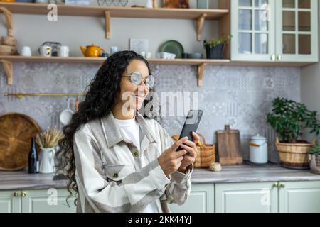 Una giovane bella donna latino-americana sta in piedi in cucina a casa, utilizzando un telefono. Sorridendo, scrive i messaggi, legge le notizie, fa le telefonate. Foto Stock