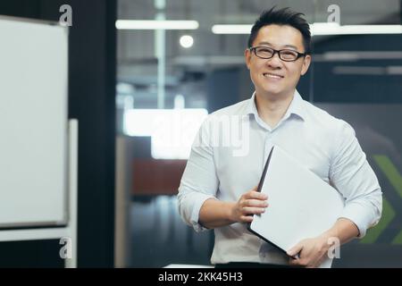 Ritratto di un bel giovane allievo asiatico sorridente. È in piedi nell'ufficio del campus indossando gli occhiali e una camicia bianca, tenendo una cartella con i documenti nelle sue mani. Foto Stock