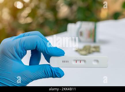 Mano con guanti che tengono una striscia positiva di risultati del test rapido dell'antigene Covid-19 e una capsula medica di blur herb. Foto Stock