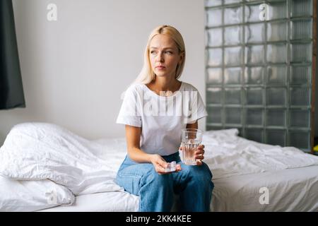 Ritratto di dubbia bionda giovane donna tenendo pillole e bicchiere d'acqua nelle mani, si sente incerto prendere la medicina. Foto Stock